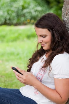 Young relaxed woman sending a text with her cellphone while sitting against a tree