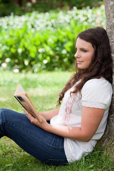 Young thoughtful girl sitting against a tree while reading a book in a public garden