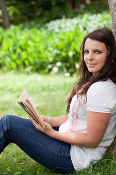 Young woman looking at the camera while leaning against a tree and holding a book