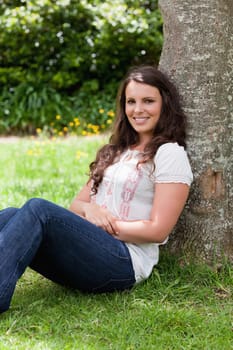 Young woman sitting against a tree while crossing her arms and beaming