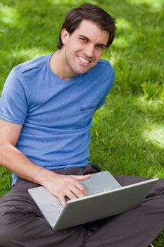 Young smiling man working on his laptop while sitting cross-legged in a parkland