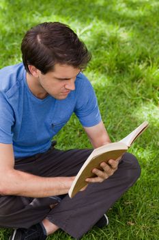 Young serious man sitting cross-legged on the grass in a park while reading a book