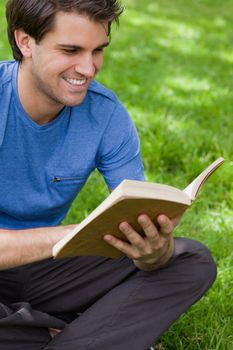 Young smiling man reading a book while siting cross-legged on the grass in a park