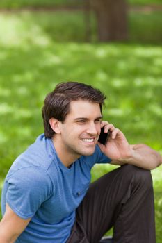 Young smiling man talking on the phone while sitting on the grass in a public garden