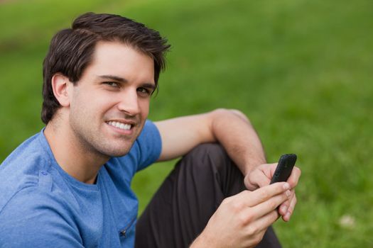 Happy young man looking at the camera while sending a text and sitting on the grass