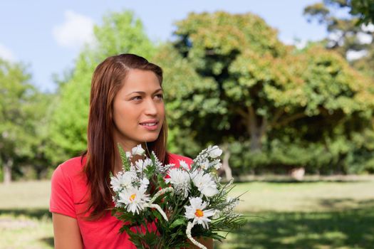 Young woman looking thoughtfully towards the side while holding a bunch of flowers in a bright park