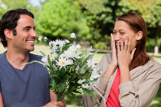 Young woman holding her hands against her face ecstatically as presented with flowers by her friend in a sunny park environment