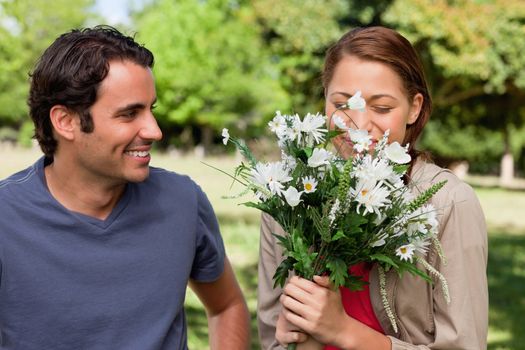 Man happily smiling as he watches his friend smell a bunch of flowers in a bright grassland area