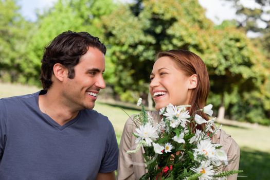 Man smiling as he watches his friend laughing while holding a bunch of flowers while sitting in a bright park