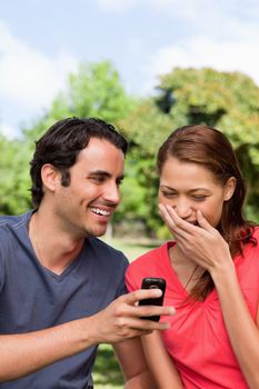 Woman covering her mouth as she is laughing at something being shown to her on her friend's mobile phone in a bright parkland