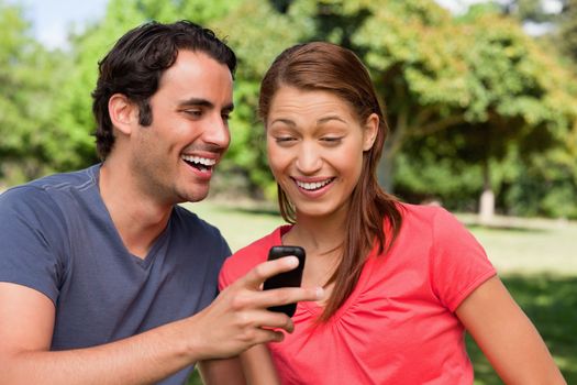 Man laughing as he shows something on his phone to his friend who is laughing happily while sitting in a bright grassland