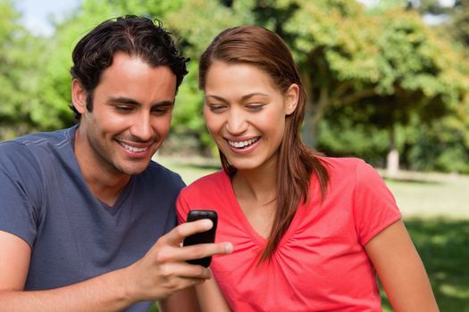 Two friends smiling as they are looking at something on a mobile phone while sitting in a bright grassland area