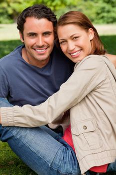 Two friends smiling and looking in front of them as they sit next to each other on the grass in a bright parkland