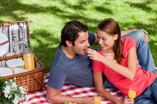 Woman putting food into her friends mouth as they lie on a blanket with a picnic basket,food and flowers