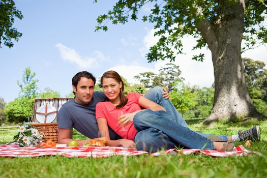 Two friends looking ahead while they hold glasses as they lie on a blanket with a view of the sky and a tree in the background