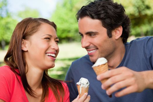 Two friends laughing while looking at each other and holding ice cream in a sunny park