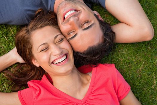 Two friends smiling with their eyes closed while lying head to shoulder with an arm behind their head on the grass