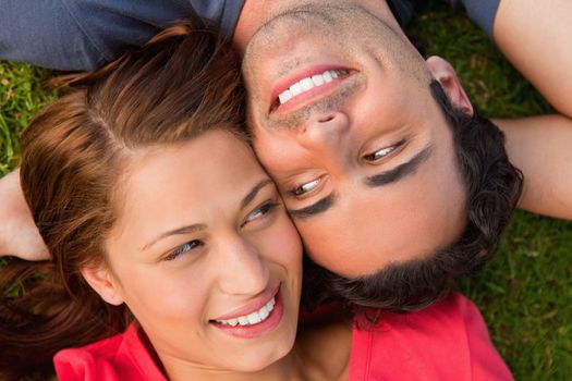 Close-up of two friends smiling while looking at each other as they lie head to shoulder with an arm behind their head on the grass