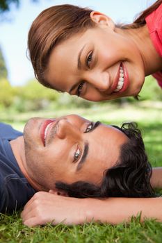 Smiling woman looking towards the side while her head is above her smiling friend's head as he is lying on the grass