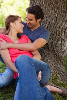 Man looking at his smiling friend as he holds her while they sit together against the trunk of a tree