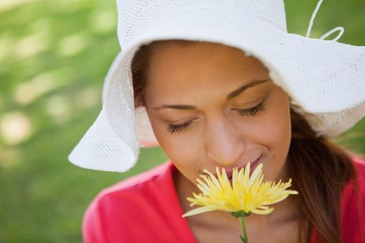 Woman wearing a white hat while smelling a yellow flower with her eyes closed, with grass in the background