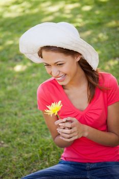 Smiling woman wearing a white hat while holding a yellow flower as she sits in the grass