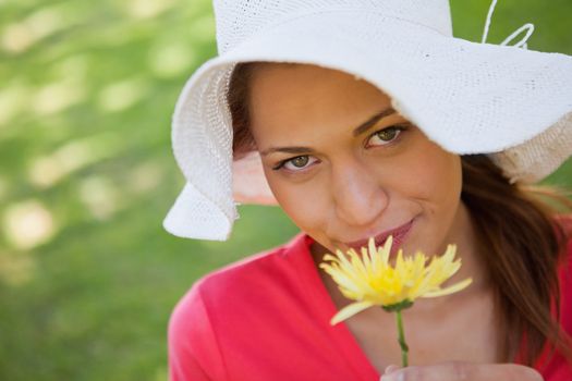 Woman looking ahead while wearing a white hat and smelling a yellow flower, with grass in the background