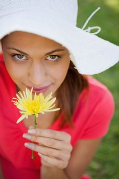 Woman wearing a white hat while smelling a flower while looking towards the camera
