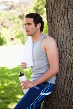 Man with a white towel on his shoulder, holding a sports bottle while leaning against the trunk of a tree