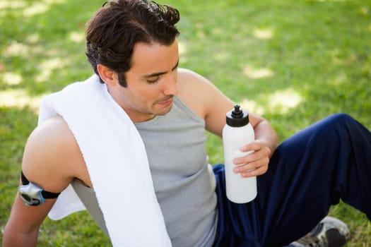 Man with a white towel resting on his shoulder looking at a sports bottle while he is sitting on the grass