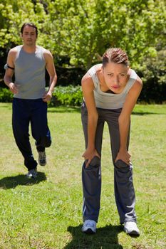 Woman bending over to recover while a man is jogging close to her in the background