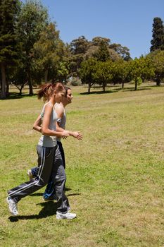 Two friends jogging along side by side other across grass with a view of trees and the sky in the background