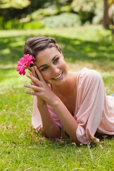 Smiling woman with her head tilted to the side lying on the grass while holding a pink flower against her head