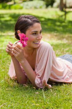 Smiling woman lying down in grass while looking towards the side and holding a flower in her hand