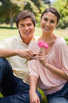 Man giving a flower to a woman as they both look straight in front of them while they sit on the grass