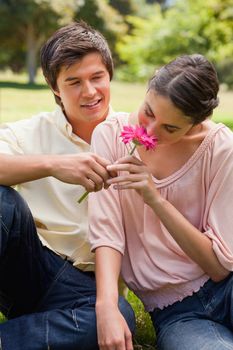 Woman smelling a pink flower which is being given to her by a smiling man as they are sitting down on the grass