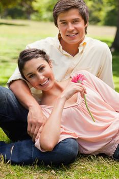 Woman smiling while holding a pink flower as she lies against her smiling friend who is sitting down on the grass