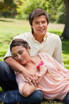 Woman with her eyes closed while holding a flower as she rests against her friend who is sitting in the grass