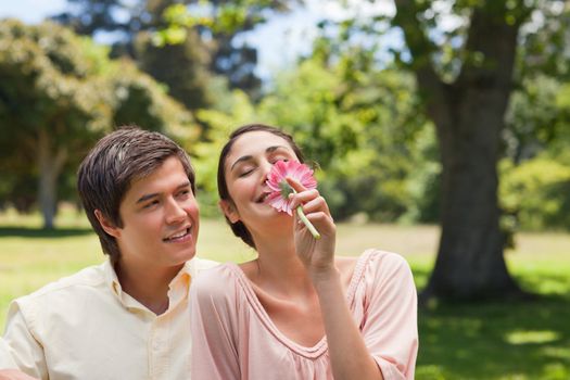 Smiling man watching his friend while she is smelling a pink flower as they are both sitting down on the grass