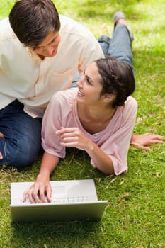 Two friends looking at each other as they are lying down on the grass with a laptop open in front of them
