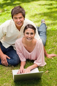 Two friends looking ahead while lying down in the grass with a laptop on the ground in front of them