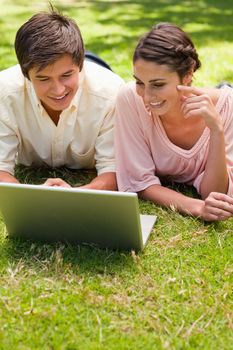 Two friends smiling while using a laptop together as they lie down in the grass