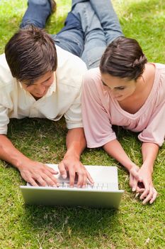 Two friends smiling while watching something on a laptop as they lie down together in the grass 