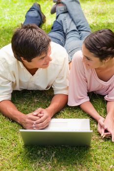 Two smiling friends looking at each other as they lie down in the grass with a laptop in front of them