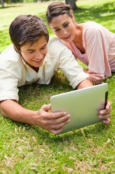 Two friends smiling as they watch something on a tablet together while lying down on the grass