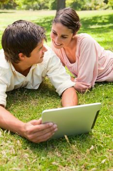Two friends smiling while looking at each other as they use a tablet together on the grass