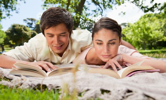 Man and woman looking down at books while lying prone on a grey blanket in the grass