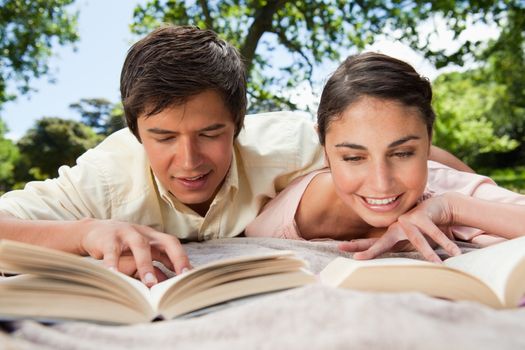 Man and a woman smiling while reading books together as they are lying prone on a blanket in the grass