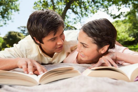 Man and a woman looking at each other while reading books as they lie prone on a grey blanket in the grass