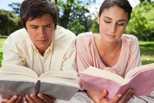 Man and a woman with a serious expression while reading books as they lie prone on a blanket in the grass